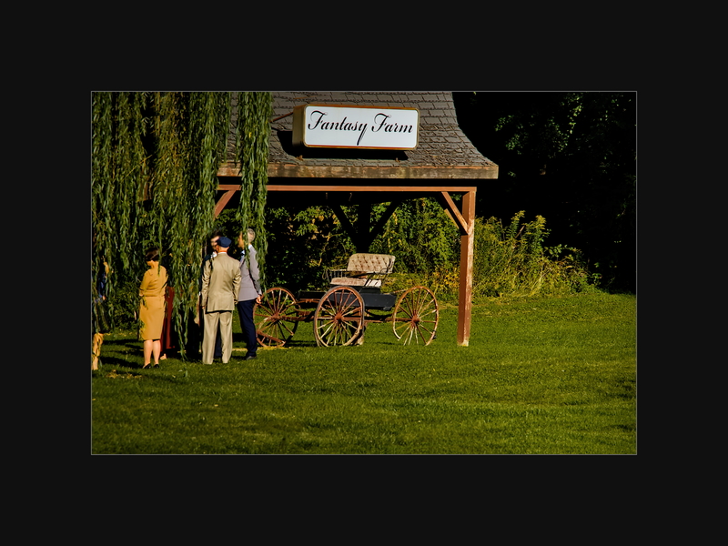 Old buggy under a shed with a FANTASY FARM sign, guests in formal dress at the left edge.